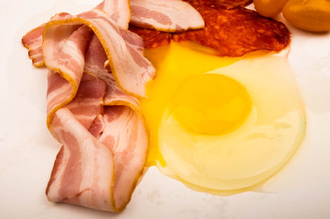 A broken chicken egg without a shell, slices of sausage and bacon and tomatoes on a white background. Close up.