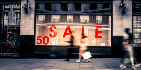 Motion blurred shoppers walking past shop window with 'Sale' sign