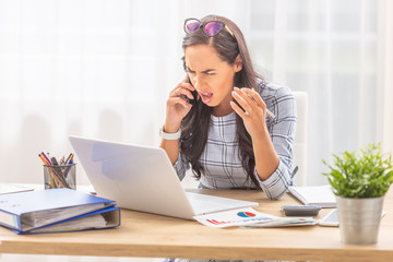 Upset businesswoman screaming into the mobile phone gesturing, while sitting in front of her computer in the office