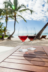 Red wine glasses with sun shadows on the wooden table standing outside at the sandy  beach with seaview on the ocean, palms and blue sky 