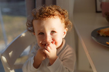 A little blond-haired curly girl in harsh sunlight sits at a table with her mouth in her hands and laughs