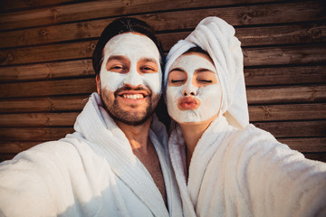 Close up selfie of beautiful happy couple after shower with a towel on head sending air kiss with cream mask on face.Woman with clay mask taking selfie with mobile phone at home enjoying relaxation.