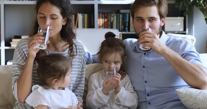 Healthy family young adult parents with small kids drinking clean fresh pure water together looking at camera at home. Happy couple and cute little children health care hydration concept. Portrait