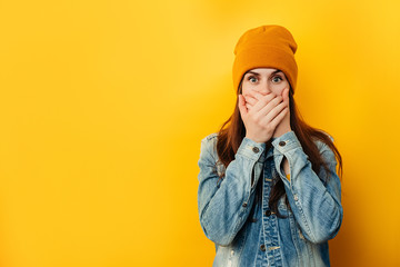Embarrassed emotional astound woman in hat looks with frightened neurotic expression directly at camera, covers mouth with both hands, dressed in denim jacket, isolated over red background