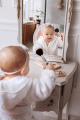 A cute little girl in a bathrobe is holding a makeup brush, doing make up and looking in the mirror at home. baby Girl is sitting on the chair near the classic mirror . Kid's fashion.