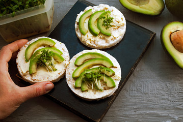 Gluten-free cakes made from rice crackers, avocados and microgreens. Female hand takes a sandwich. Black cutting board. Vegan breakfast rice tortillas avocado.