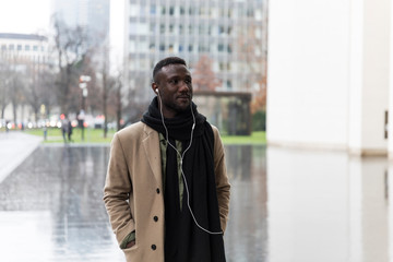 Young Black Man in Beige Coat and Earphones Walking in City