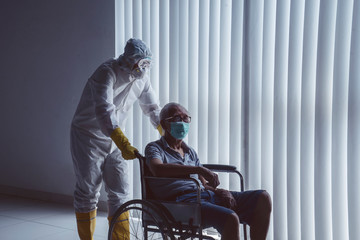Doctor with hazardous materials suit pushing an aged patient in wheel chair infected with coronavirus