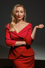 Fototapeta premium Blonde woman with bright make-up and deep neckline, in red dress and black earrings. She smiling, posing on gray background. Copy space, close up