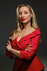 Blonde girl, bright make-up and deep neckline, hands folded, in red dress and black earrings. Smiling, posing sideways on gray background. Close up