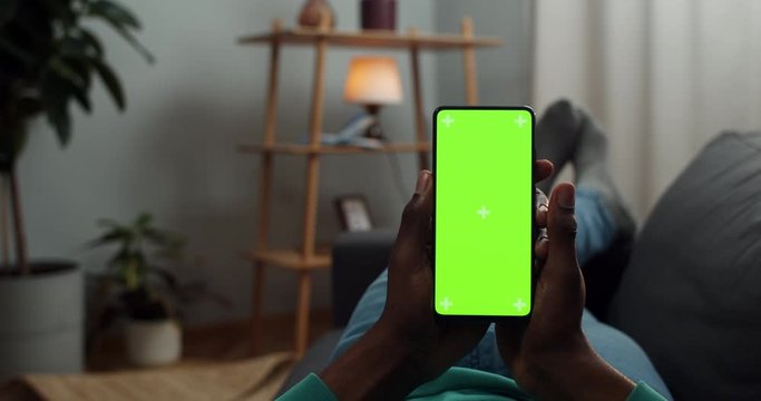 Afro american male person lying on couch and holding modern phone with both hands. Man browsing internet while looking at mock up screen with trecking markers . Concept of chroma key.