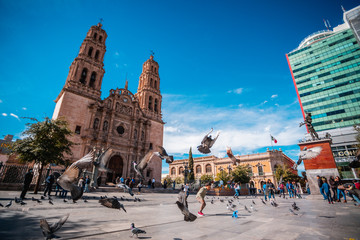 Plaza pública frente a una iglesia con aves volando y personas jugando.