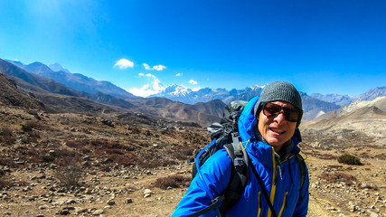 Naklejka premium A man hiking through dry path in Himalayan valley, located in Mustang region, Annapurna Circuit Trek in Nepal and taking a selfie. The man carries a heavy backpack. Barren slopes. Harsh landscape.