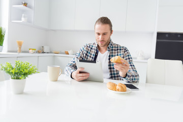 Portrait of focused guy sit kitchen table use tablet watch film video have snack eat tasty delicious yummy croissant wear casual checkered shirt in house indoors