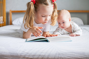 A caring older sister is reading a book to her younger newborn sister while lying on the bed. It's good to be home.