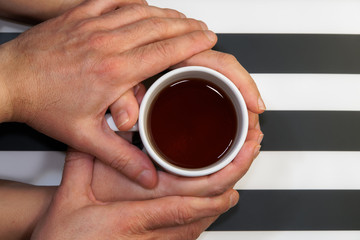 Loving couple on relationship, affectionately holding hands over beverage cup top view. Close up on male tenderly touching female hands that hold a tea mug at home.