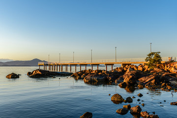 tourist pier under blue sky in calm sea with rocks in the foreground and mountains on the horizon - 343583539