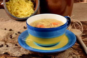 Soup with chicken noodles in a blue mug next to a clay pot, from which the cooked noodles are poured on the table
