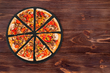 Sliced pizza with mushrooms, corn, cherry tomatos, courgettes and bell peppers on a slate round platter on the brown wooden background, top view