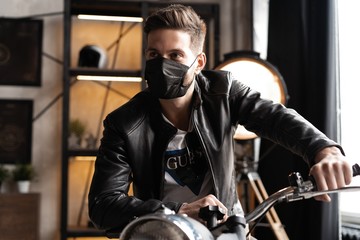 Handsome brutal male biker in black mask in leather jacket sitting on motorcycle looking forward.