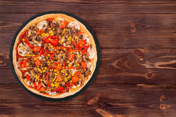 Delicious whole pizza with mushrooms, corn, cherry tomatos, courgettes and bell peppers on a slate plate on the wooden table, top view