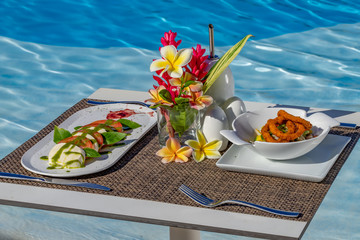 Caprese salad and fried squids on a white plate, fork and knife, glass of water, restaurant table by a pool, caribbean flowers, blue water background