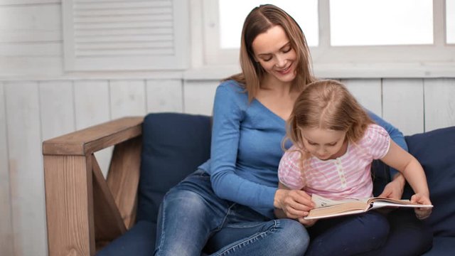 Caring young mother and cute girl reading book together sitting on couch. Shot with RED camera in 4K