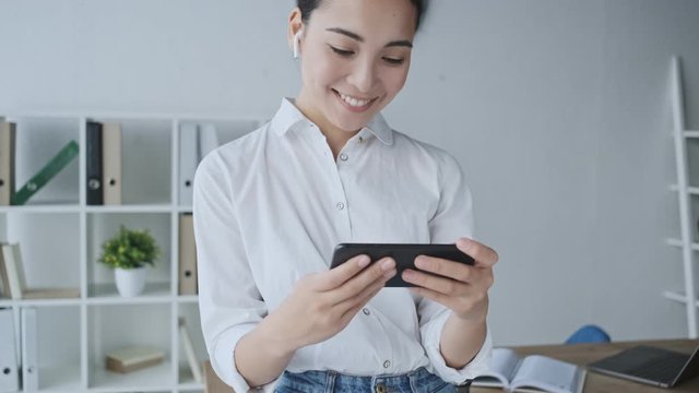 An attractive young asian businesswoman with wireless earphones is watching something on her mobile phone while sitting in the office