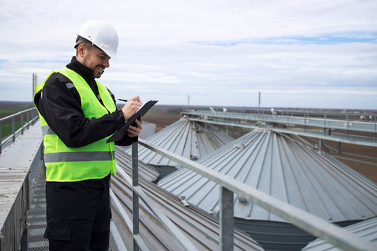 Portrait Of Construction Worker Standing On Rooftops Of High Silos Storage Tanks And Working On Tablet Computer.