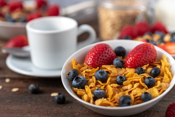 Cornflakes with blueberries and raspberries in white bowl on dark wooden desk.
