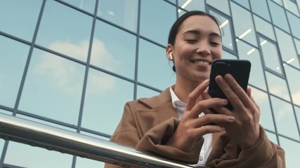 A bottom view of a beautiful pleased young asian businesswoman with earbuds is using her smartphone standing near modern office buildings