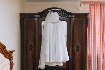 Wedding white long dress of the bride hangs on a hanger, on a wooden wardrobe with a mirror in the hotel room. Photography, concept.