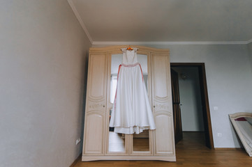 Wedding white long dress of the bride hangs on a hanger, on a wooden wardrobe with a mirror in the hotel room. Photography, concept.