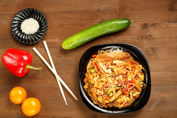 Black plate with funchose and chicken with vegetables on a wooden table and fresh vegetables.