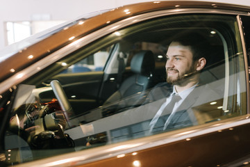 Portrait of confident bearded businessman wearing business suit sitting behind wheel of new car in auto dealership, view from window. Concept of choosing and buying new car at showroom