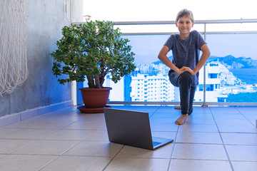 Kid with laptop computer doing sport exercises at home on balcony. Sport, healhty lifestyle, active leisure, stay at home, online learning, online training