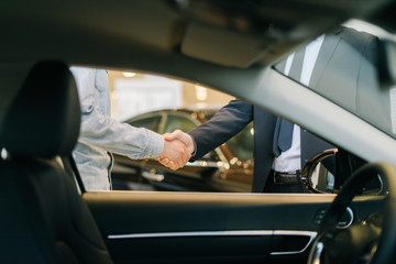 Buyer of car shaking hands with seller in auto dealership, view from interior of car. Close-up of...