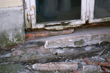 Part of the white wooden frame of an old window with peeling paint and rotten wood and plaster on facade crumbles due to moisture and mold