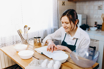 Happy young woman in apron sitting at wooden table looking to the notebook with recipes  in a kitchen. Cooking at home concept.