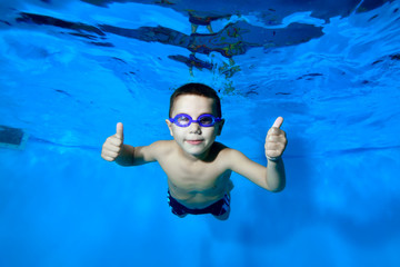 A sporty little boy poses for the camera underwater in a children's pool wearing swimming glasses. He looks at the camera and gives a thumbs-up. Closeup. Concept. Digital photo. Horizontal view. © alexbard