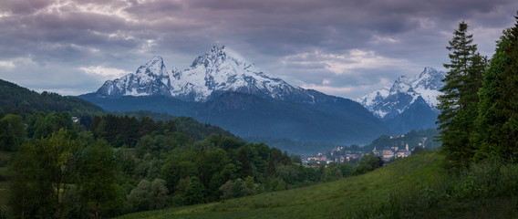 Naklejka na ściany i meble Snow covered mountain peak panorama of Watzmann and city Berchtesgaden with clouds during sunrise, green forest and fields in foreground, Bavaria.