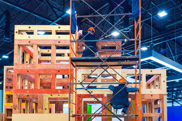 Workers on scaffolding mount a wooden frame. Installation of an advertising element in the exhibition hall. Preparation for the exhibition. Construction work.