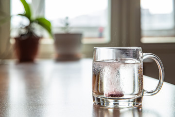 Sparkling tablet throw into a glass transparent glass, the glass is on a table, a pill for cold of claret color, bubbles rise up, splashes take off from a glass, flowers is on background, foam