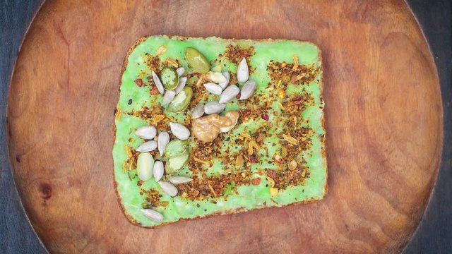 Cooking avocado toast with pepper spices, walnuts, seed of pumpkin and sunflower. Stop motion animation. Healthy food vegan background. Natural wooden plate, top view.