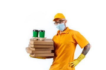 A cautious young man from a courier delivery service in a medical mask and gloves holds in his hands boxes of pizza and cups of coffee on a white background