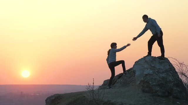 Man and woman hikers helping each other to climb stone at sunset in mountains. Couple climbing on high rock in evening nature. Tourism, traveling and healthy lifestyle concept.