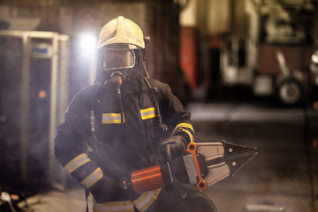 Obraz premium firefighter portrait wearing full equipment, oxygen mask, and power hydraulic cutting tool, smoke and fire trucks in the background.