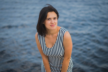 a young brunette girl of European descent in a striped dress walks along the seashore or river
