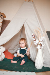 Sweet 1 year old little baby boy playing in a teepee game tent in a children's room decorated in...