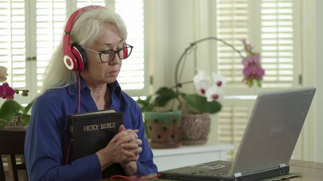 An older woman holding a Bible and praying as she attends church virtually via her laptop due to congregation and group fellowship restrictions during COVID19 pandemic.